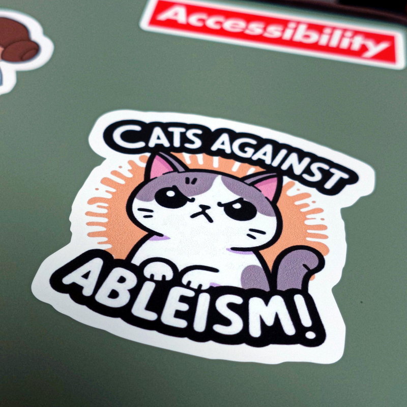憤怒貓咪圖樣搭配 "Cats Against Ableism!" 文字的貼紙貼在綠色筆記型電腦上蓋，遠處還有一張紅底白字寫著 "Accessibility" 的貼紙