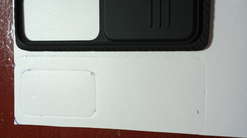 鏡頭滑蓋放在 3M VHB™雙面膠的白色雛形紙上，雛形紙有著依鏡頭滑蓋輪廓描繪出的壓痕